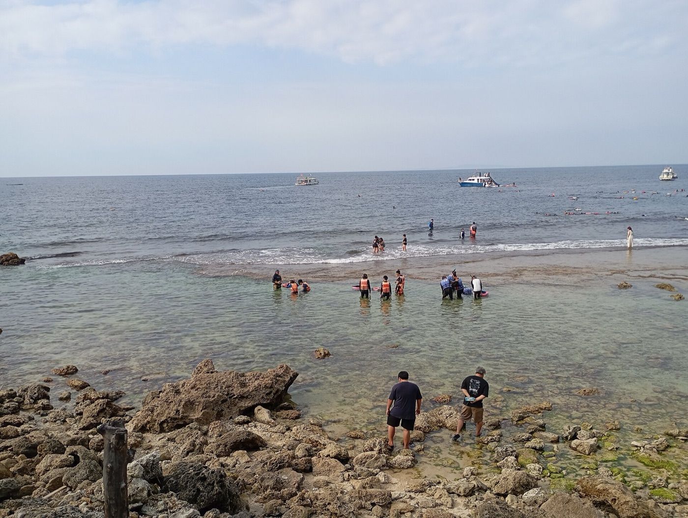 礁岩海岸邊，兩名男子踩著礁石走進淺水，前方淺灘上幾群穿救生衣的人站在水中，遠方海面停著三艘船