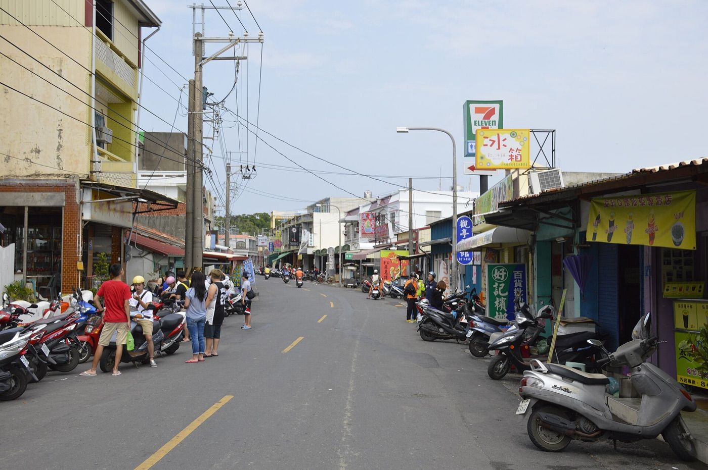 中正路街景，雙線柏油路兩側停滿機車，右側有便利商店和多面店家招牌，左側幾名遊客站在機車旁