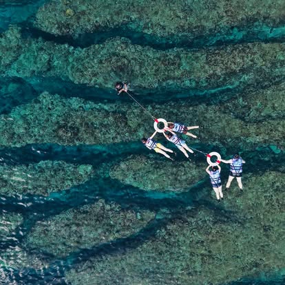 空拍視角：穿救生衣的浮潛客拉著浮圈排成隊形，浮在清澈的珊瑚礁海面上